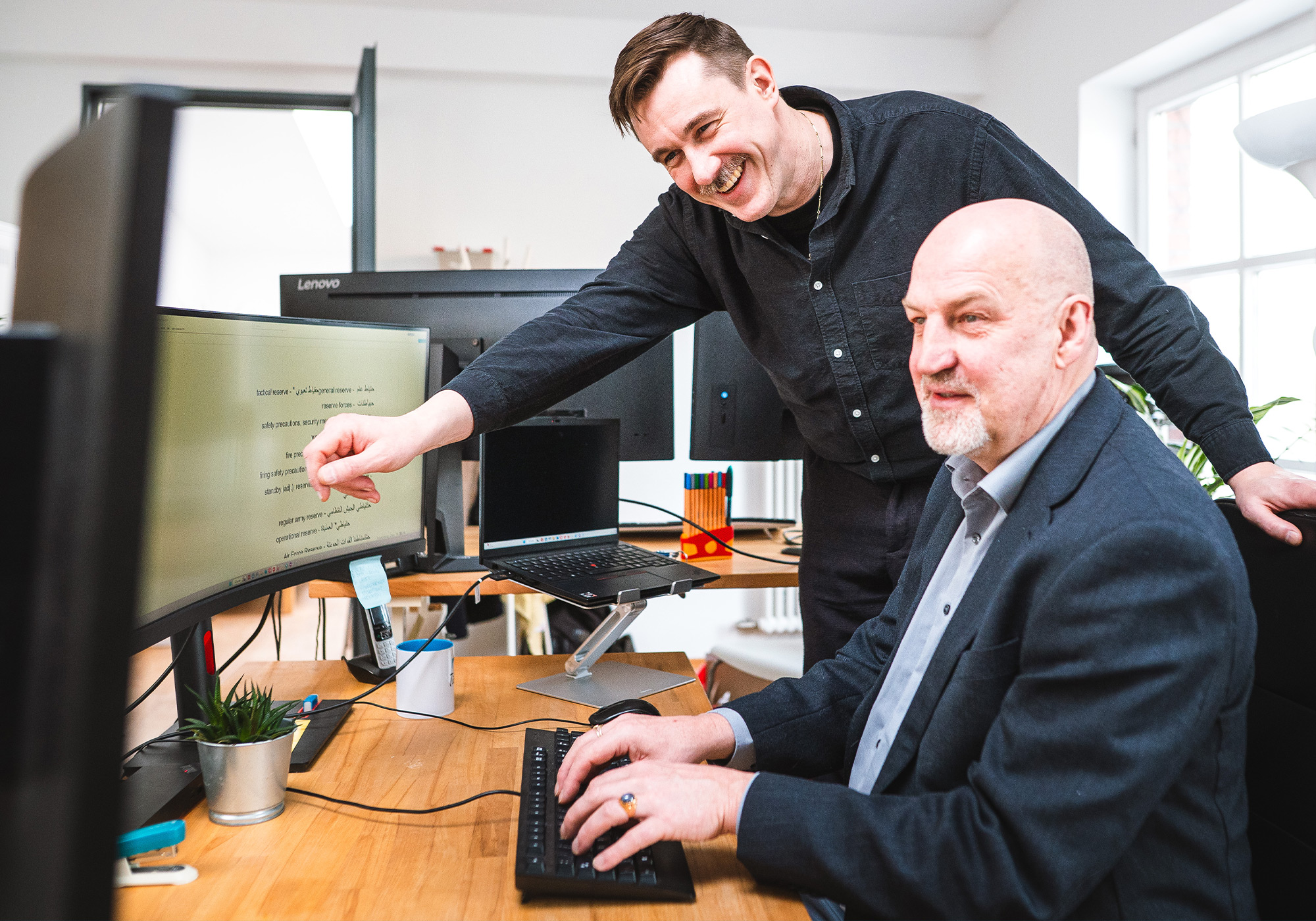 Zwei männliche Kollegen schauen auf den Desktop eines Computers und lächeln dabei