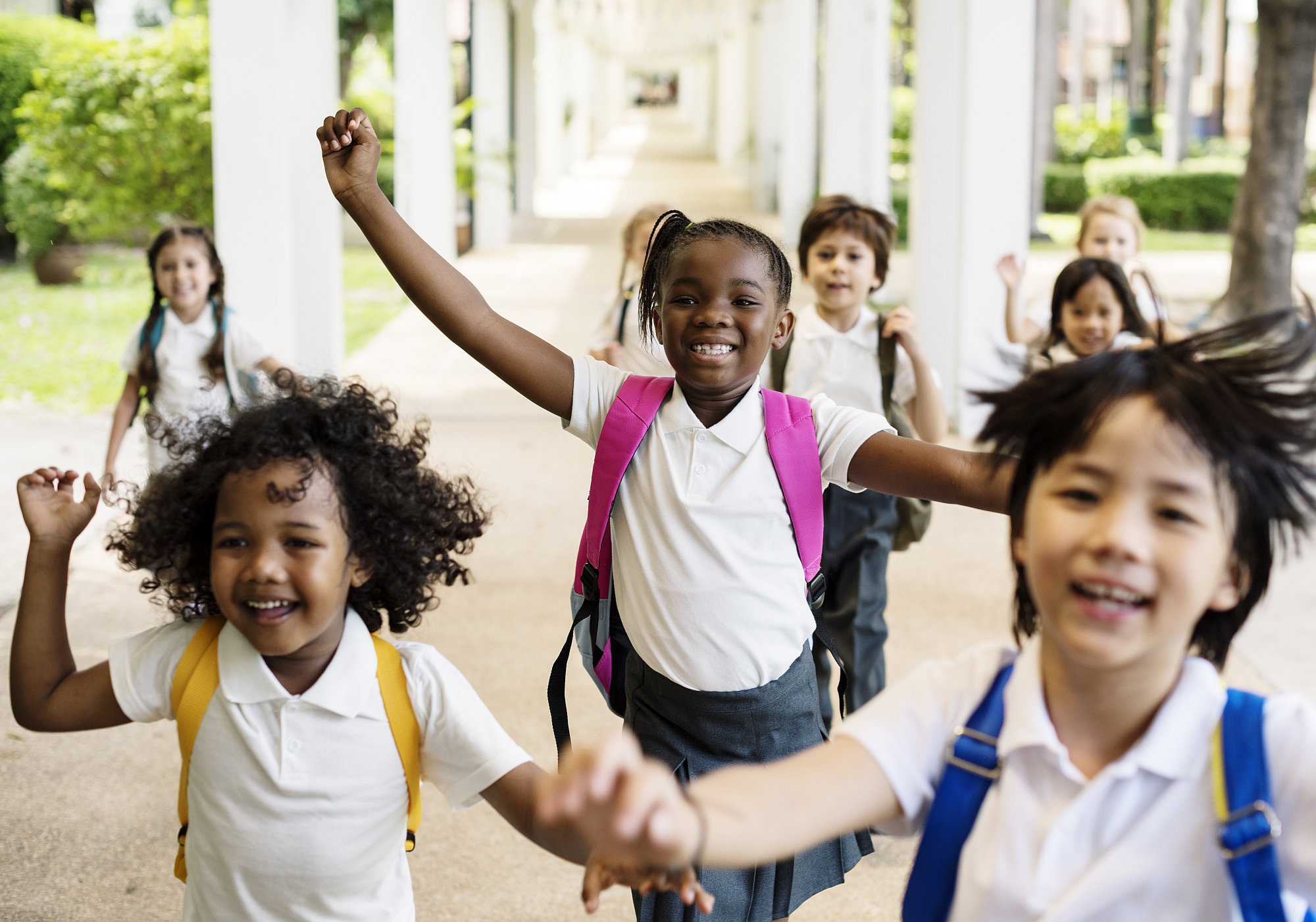 Acht internationale Grundschulkinder mit weissen T-Shirts und bunten Rucksäcken jubeln