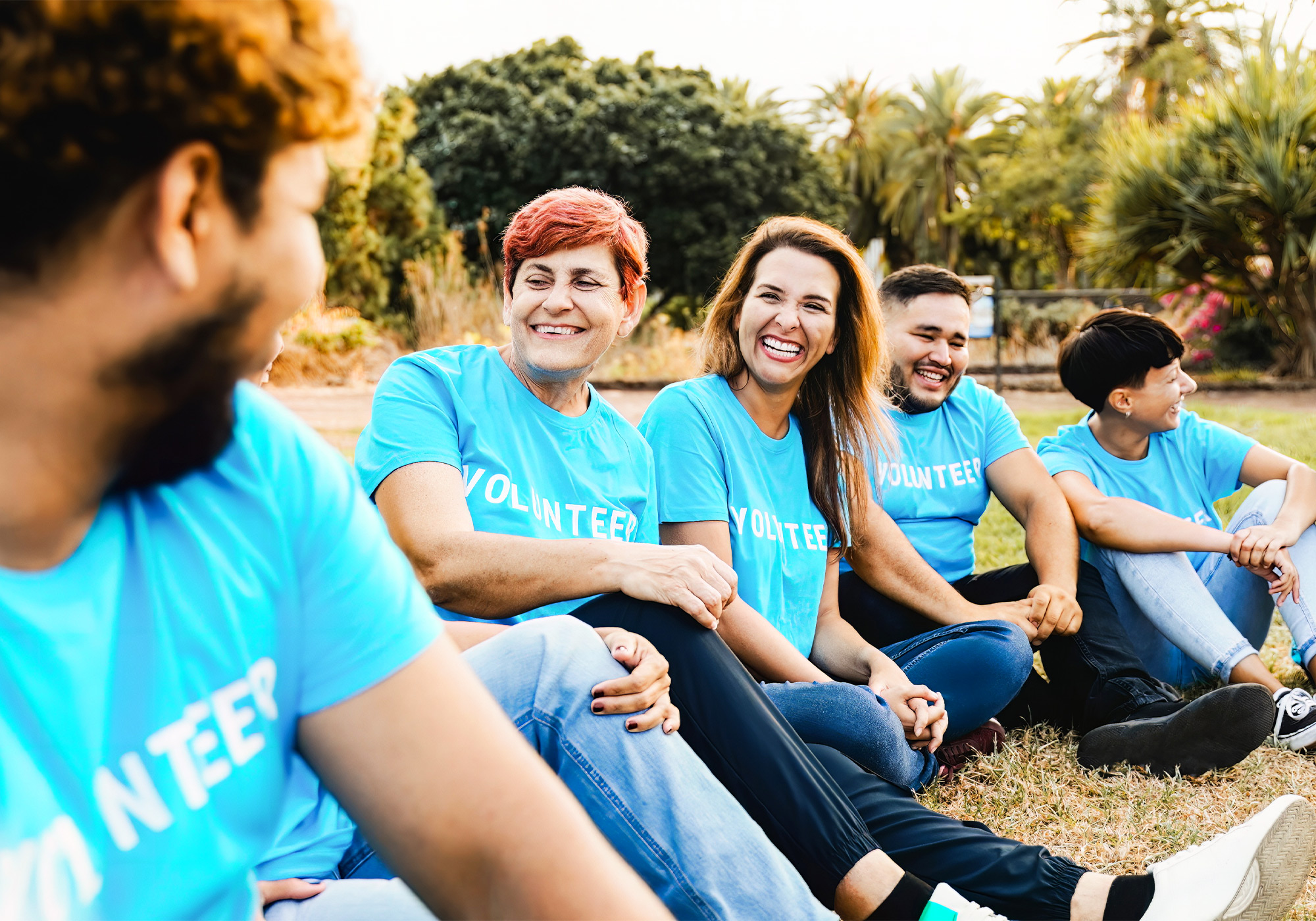 Team einer gemeinnützigen Organisation sitzt im Gras auf dem Boden und lacht. Alle tragen ein blaues T-Shirt mit de Aufschrift Volunteer.
